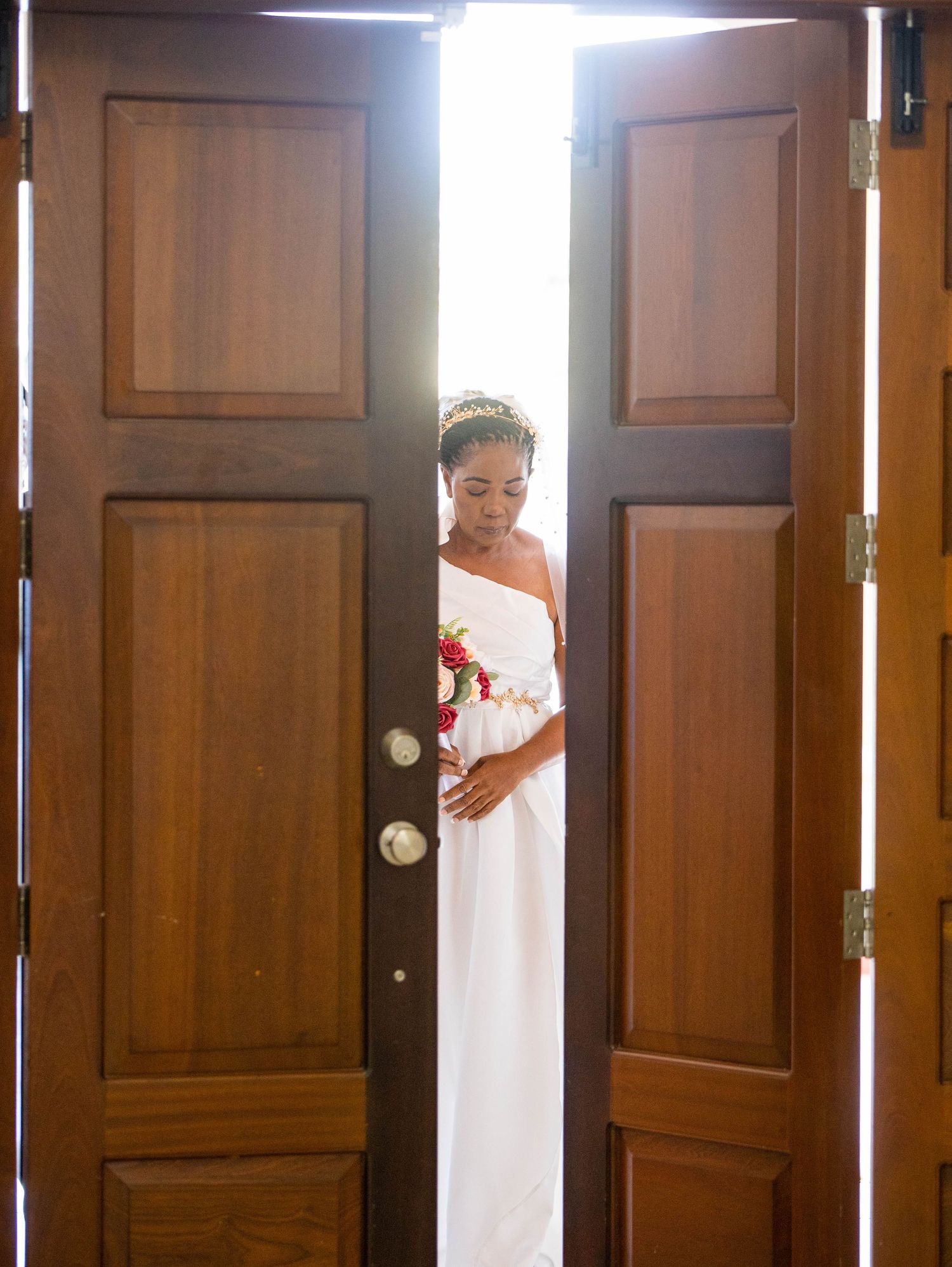 A bride in a classic white gown stands silhouetted in open wooden double doors with bright light streaming through.