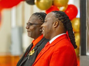 Two people in bright red suits and corsages standing together at a formal event with gold and red balloons in background.