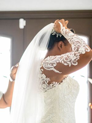 A bride wearing an intricate lace wedding dress with illusion back detail and flowing white veil adjusts her hair.