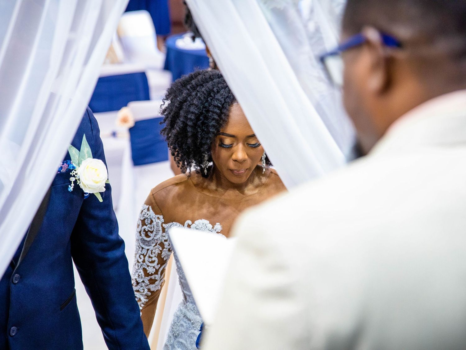A bride in an elegant off-shoulder lace wedding gown gazes down during a ceremony with royal blue decor.