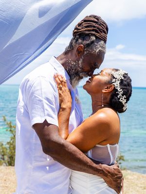 A romantic wedding kiss captured on a beautiful beach with turquoise waters and blue sky in the background.
