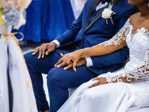 A couple in wedding attire sits close together, wearing a navy blue suit and white lace dress with floral boutonniere.