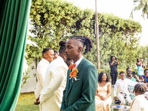 A person in an emerald green suit with an orange boutonniere stands at an outdoor garden wedding ceremony.