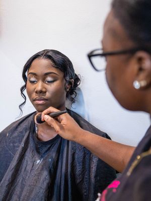 A makeup artist applies cosmetics on a client wearing a black salon cape during a professional beauty session.
