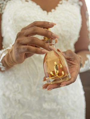 Elegant hands holding a golden perfume bottle against a white lace wedding dress background.