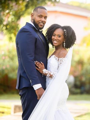 Two newlyweds pose together outdoors in formal wedding attire with a blurred garden backdrop.