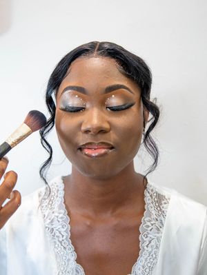 A makeup artist applies cosmetics with a brush to a bride wearing a white lace robe for wedding preparation.
