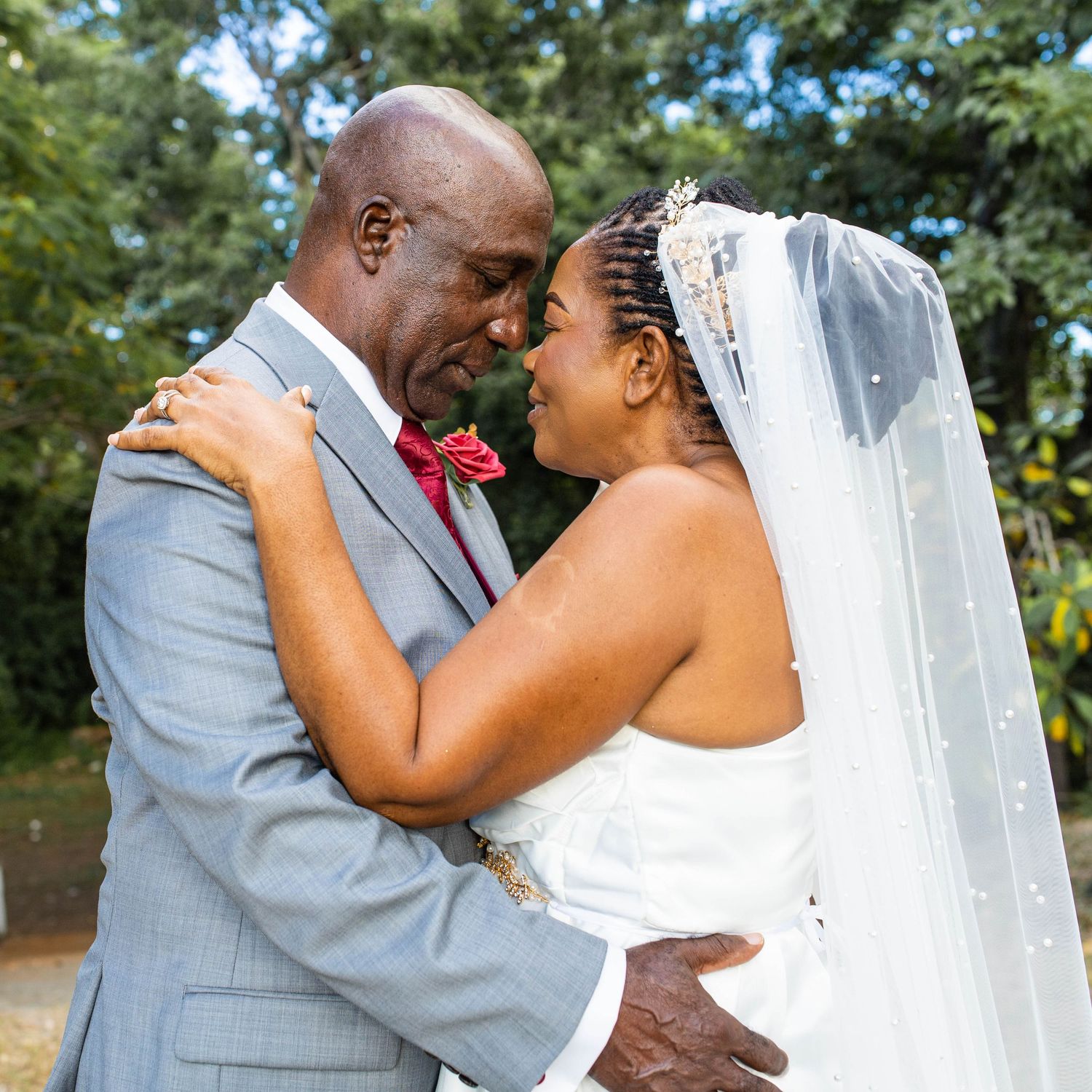 A loving bride and groom share a tender moment during their outdoor wedding, with trees and greenery in the background.