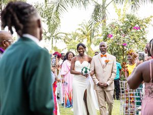 A joyous outdoor wedding ceremony takes place among palm trees and pink flowers in a tropical garden setting.