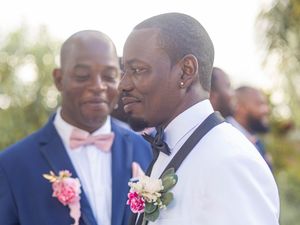Two men in formal attire with matching pink floral boutonnieres share a joyful moment at an outdoor celebration.