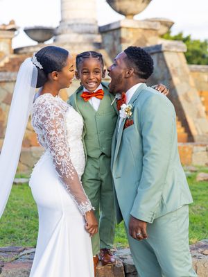 A heartwarming wedding moment showing a bride in white lace, young boy in green suit, and groom in light suit sharing a laugh.