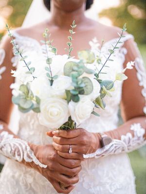 Elegant bridal bouquet with white roses and eucalyptus stems held by hands wearing delicate lace sleeves and engagement ring.