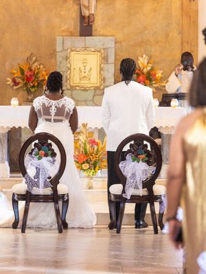 A couple at their wedding ceremony sitting in ornate chairs at altar with floral arrangements and candles in a church.