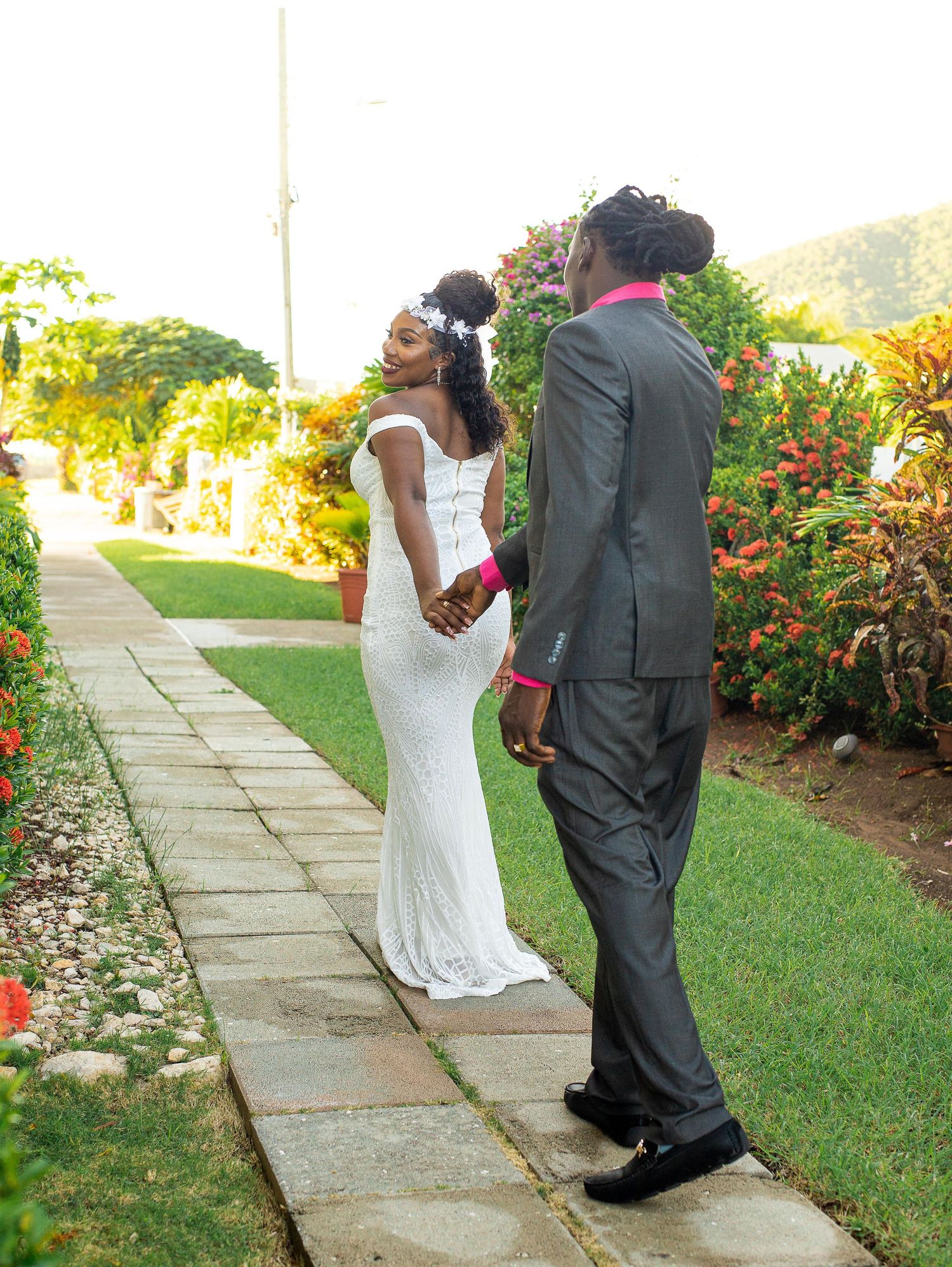 Couple in wedding attire stands on a stone path in a lush tropical garden with colorful flowers and greenery.