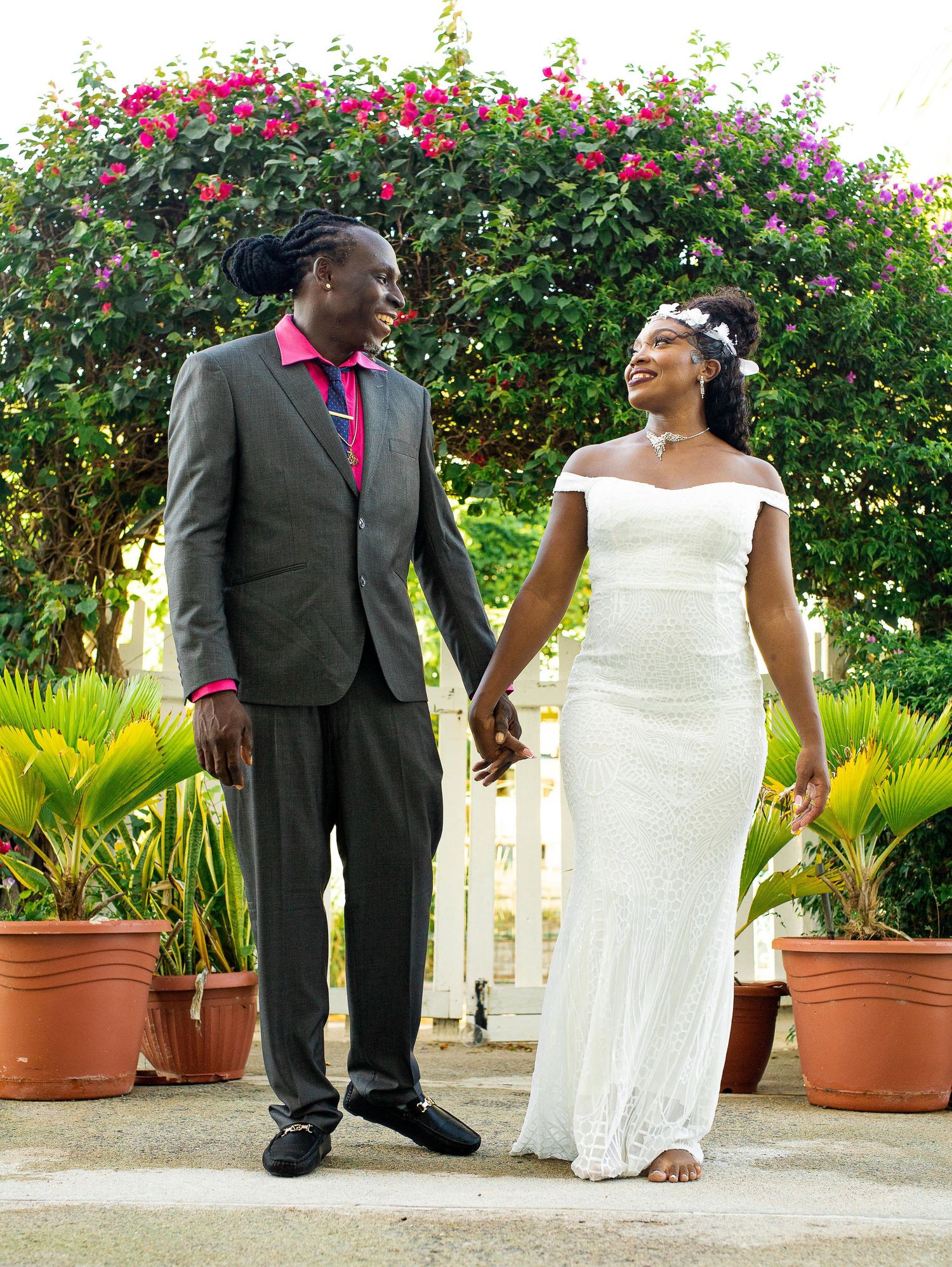 A couple in wedding attire stands together holding hands in front of a blooming floral garden with potted plants.