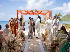 A beach wedding ceremony takes place under an orange arch decorated with flowers, with the ocean and mountains in view.
