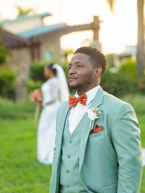 Groom wearing a mint green three-piece suit and coral bow tie stands outdoors during a wedding celebration.