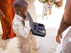 A young ring bearer in a tan suit holds a small cushion with wedding rings during a wedding ceremony.