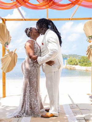 A couple shares a romantic kiss under orange draping and palm fan decor at a tropical waterfront wedding ceremony.
