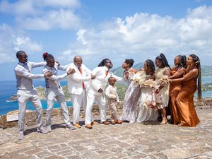 Wedding party in coordinated white and earth-tone outfits poses playfully on a coastal cliff with blue ocean backdrop.