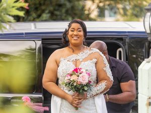A bride in a white lace wedding dress holds a pink and white bouquet while standing next to a sleek car.