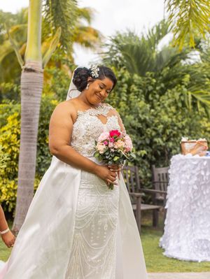 A bride in an elegant white lace wedding dress holds a pink bouquet while standing in a tropical garden setting.