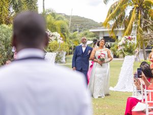 A wedding ceremony takes place outdoors in a tropical garden setting with palm trees and white chairs.