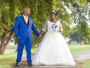 A couple in bright blue and white formal attire walking together on a tree-lined path during their wedding celebration.