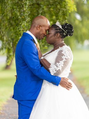 A couple in wedding attire shares a romantic embrace under weeping willow trees in an outdoor garden setting.