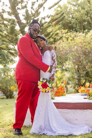 Newlyweds pose together outdoors, with bride in white lace gown and groom in bright red suit holding colorful bouquet.