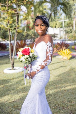 Elegant bride in long-sleeve lace mermaid wedding dress holds colorful bouquet in sunny tropical garden setting.