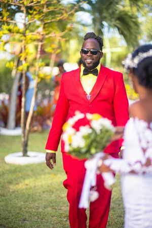 Someone in a bright red suit with bow tie and sunglasses smiles brightly during an outdoor wedding ceremony.