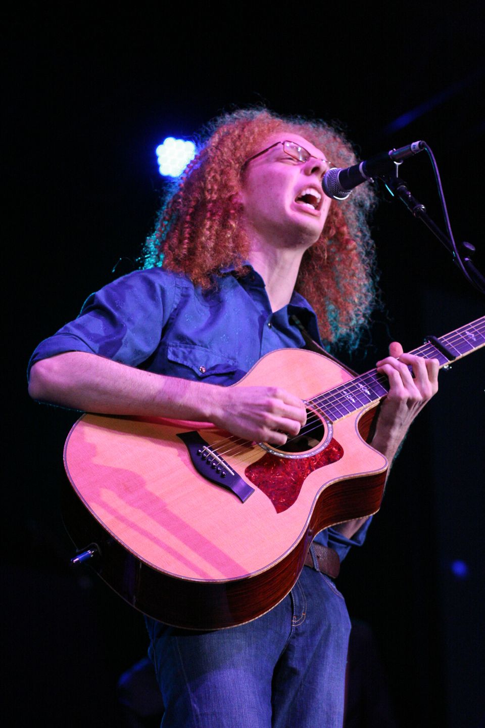 Musician with curly hair plays acoustic guitar and sings passionately on stage under blue lighting.