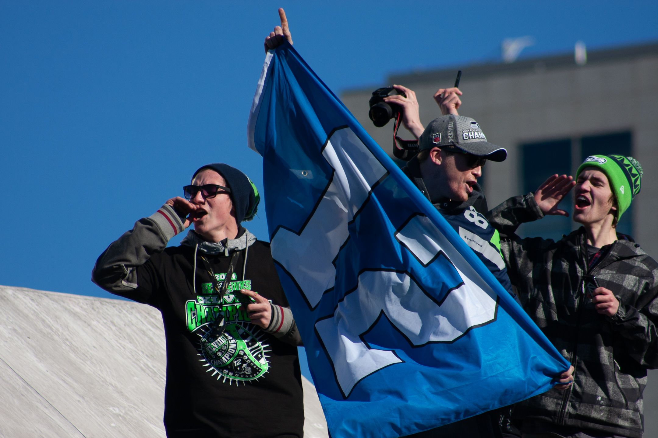 Sports fans wave a blue team flag while cheering in excitement against a bright blue sky during an outdoor event.