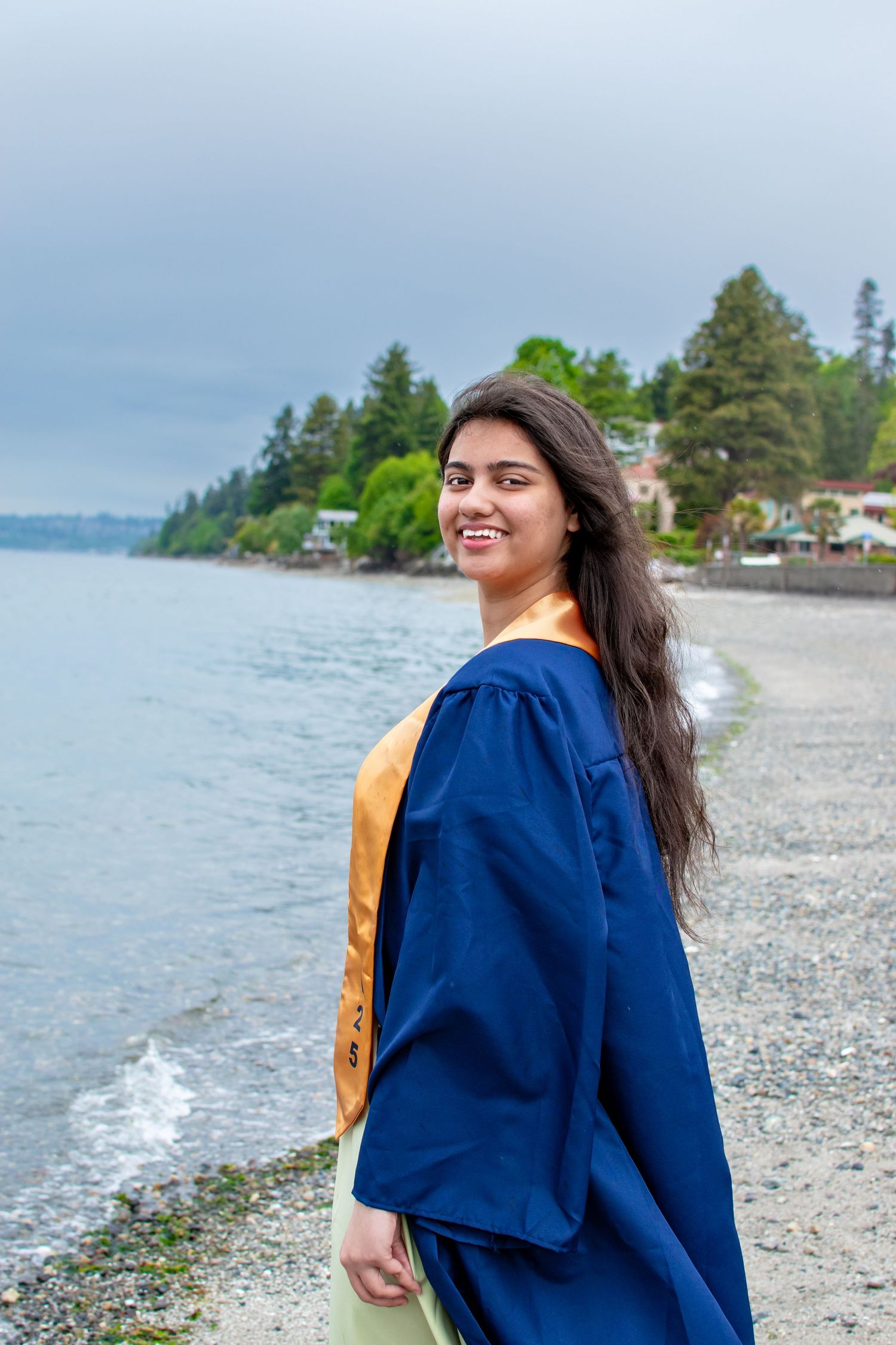 Graduate smiling over her shoulder in cap and gown along the shoreline—senior photos in Federal Way, WA.