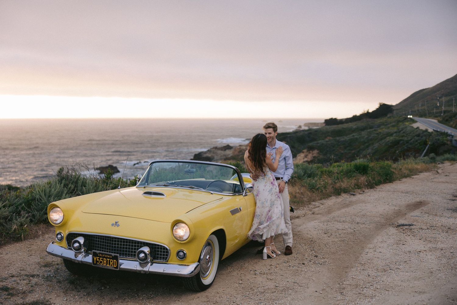 A classic yellow Ford Thunderbird parked on a coastal road at sunset overlooking the ocean.