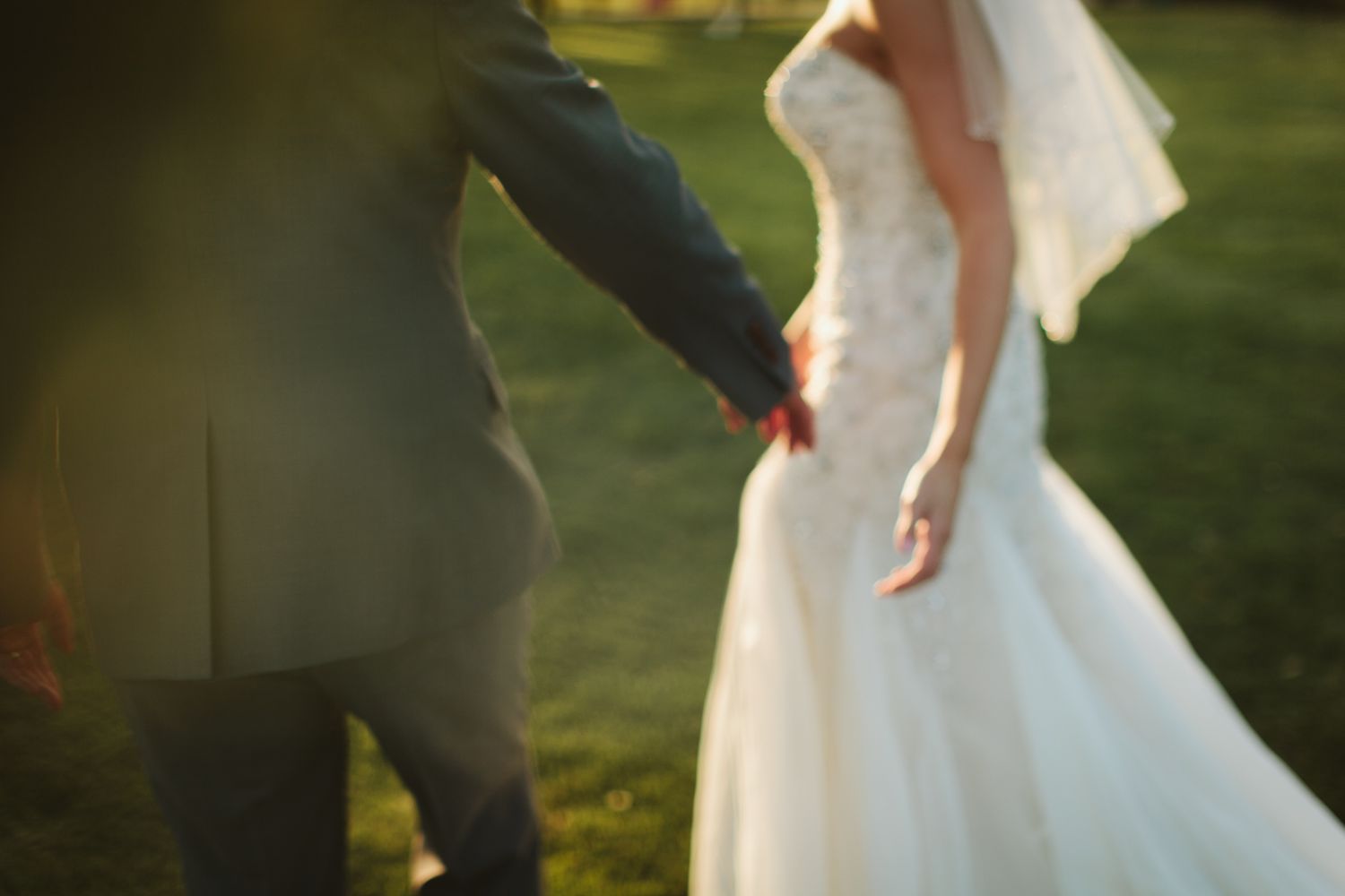 Artistic close-up of couple holding hands in wedding attire at sunset.
