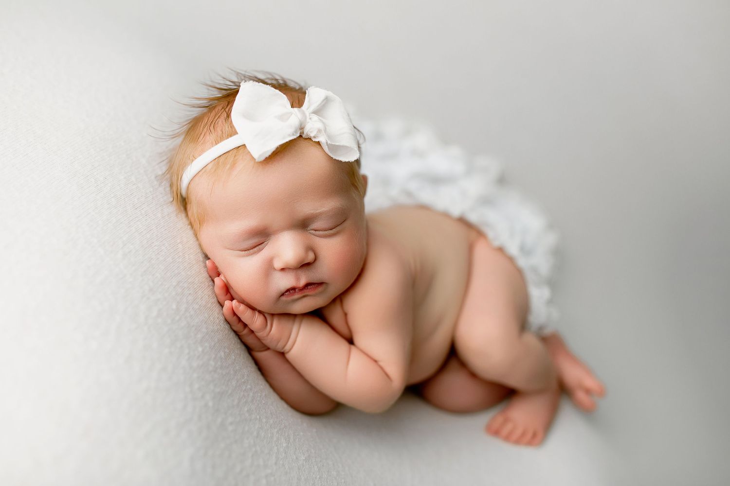 Newborn baby girl sleeping in curled pose with white bow headband on cream backdrop at Summerville SC photography studio