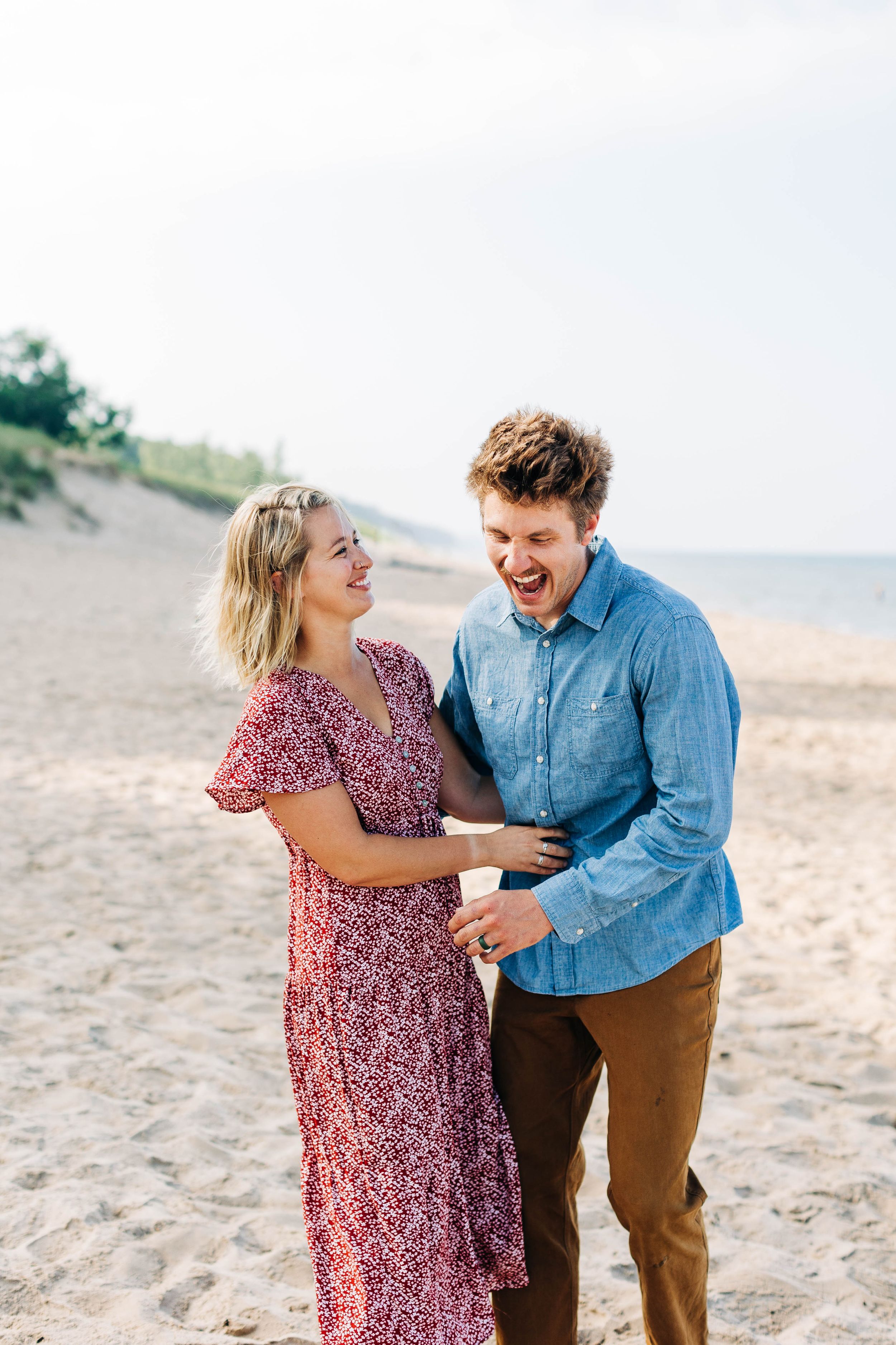A joyful couple laughing together on a sandy beach during their engagement session.