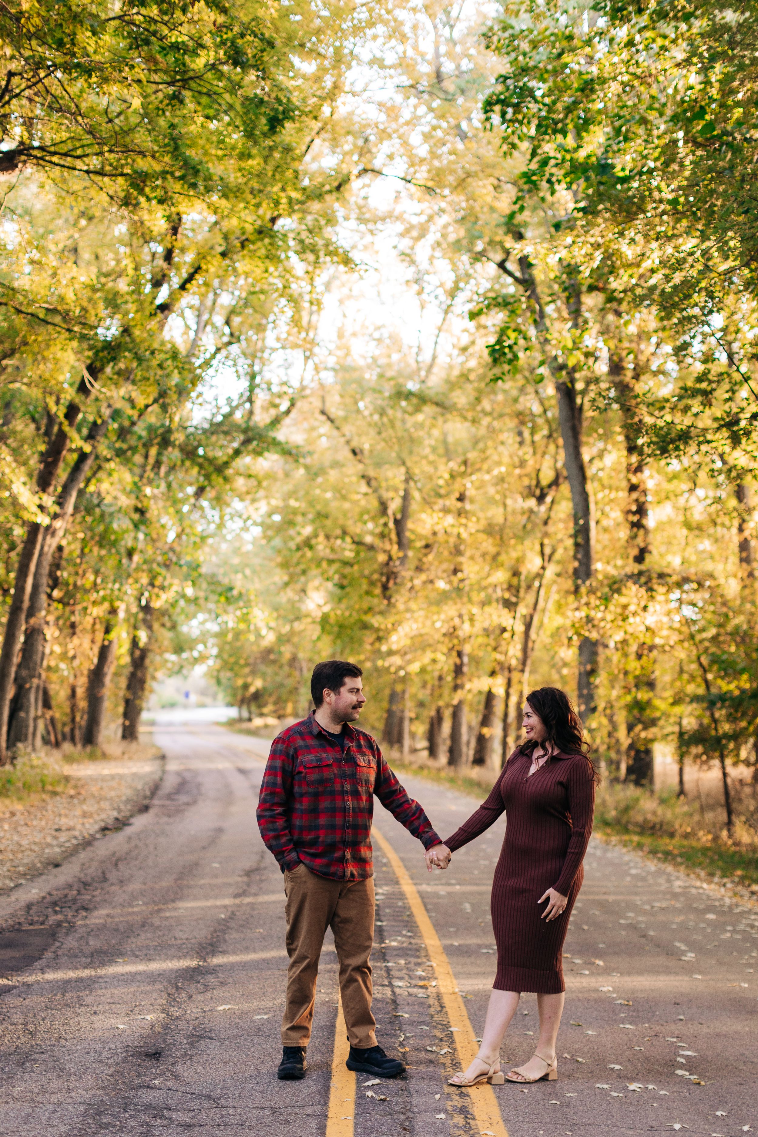 Couple holding hands on a tree-lined road with golden fall leaves and sunlight filtering through.