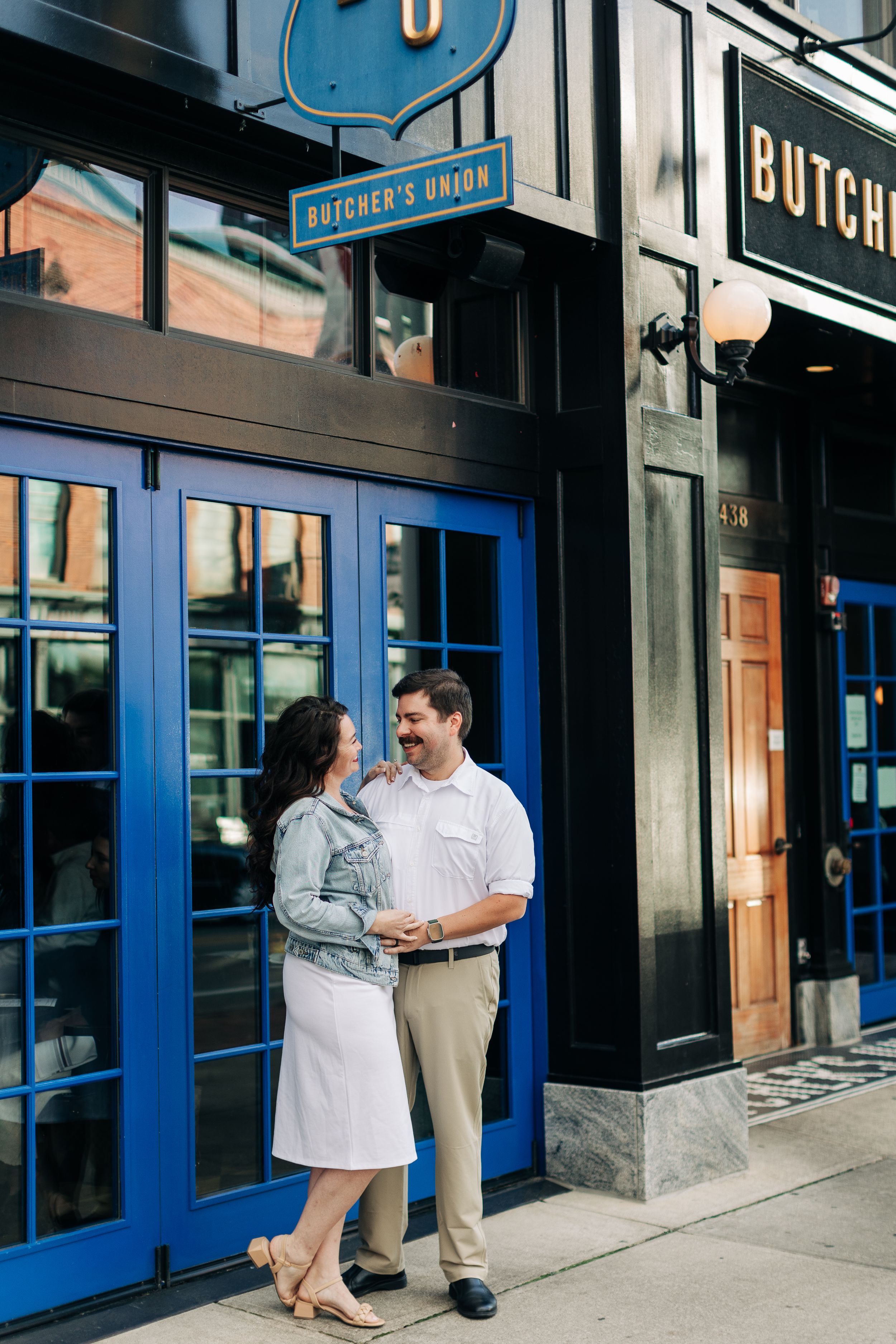 A romantic couple shares a moment outside a storefront with blue doors and vintage signage.