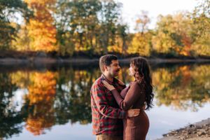 A couple embraces by a lake during autumn with vibrant fall foliage reflecting in the water.