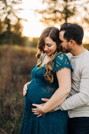Intimate maternity portrait of couple embracing in sunlit field wearing teal dress and cream sweater.