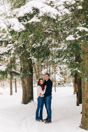 Couple embraces in snowy forest path surrounded by snow-covered evergreen trees.