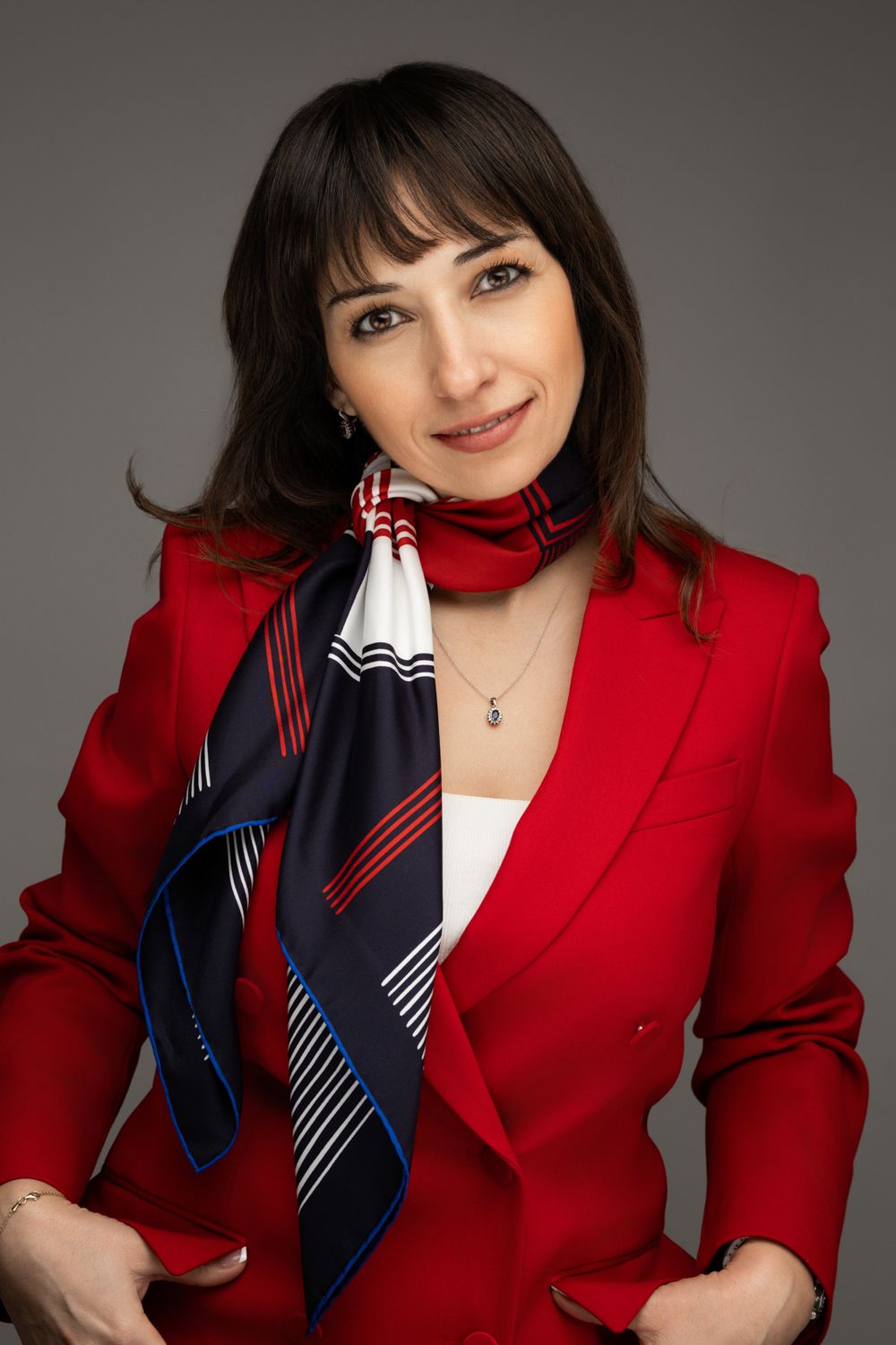 Professional portrait of someone wearing a bright red blazer and navy patterned silk scarf against a gray background.