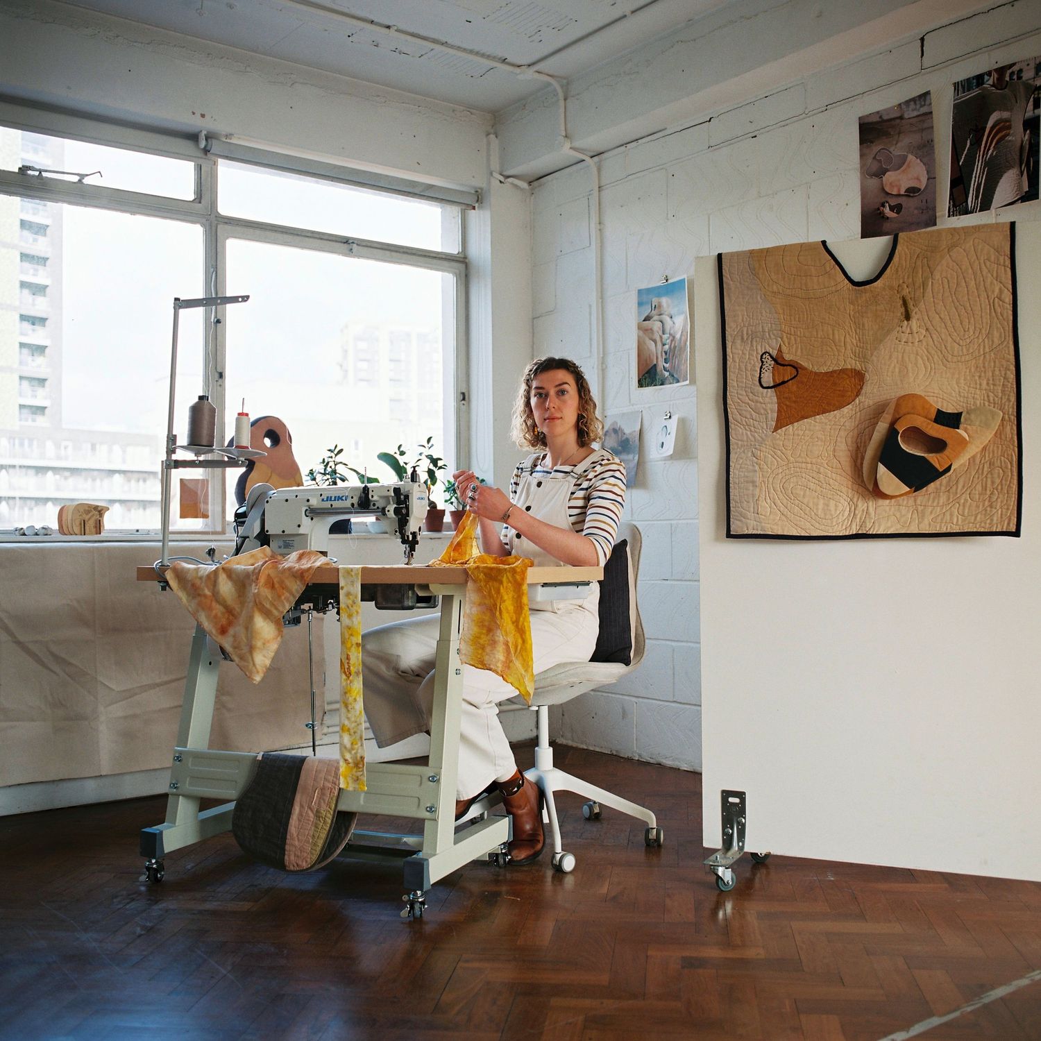 Young blonde haired women sitting at a sewing machine within her white walled studio