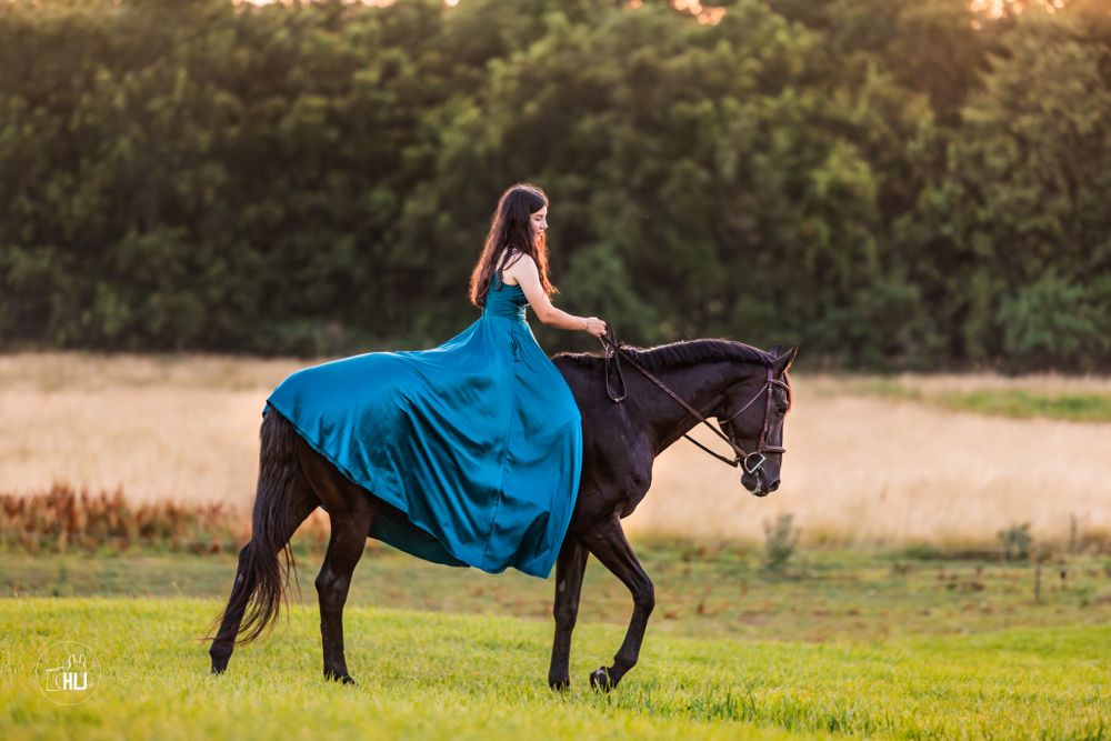 Rider in flowing turquoise dress riding elegant black horse through meadow at sunset.