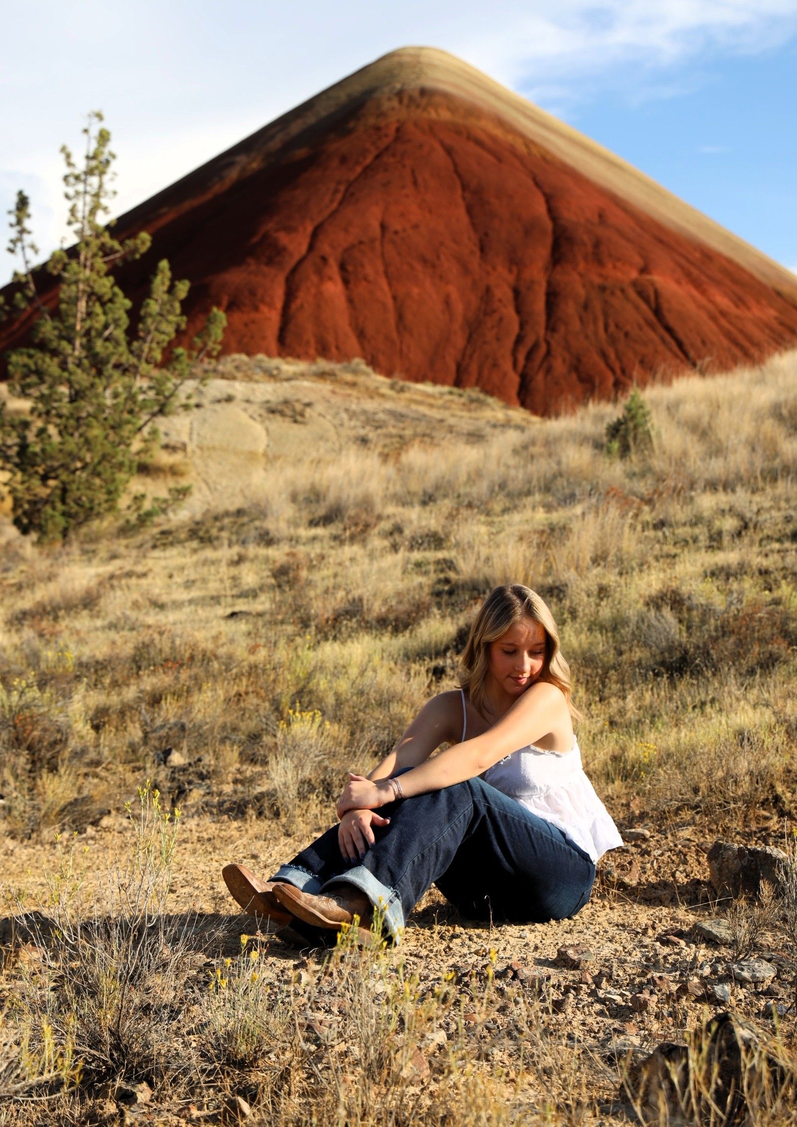 A person lies on a grassy hillside with red-colored Painted Hills formations visible in the background during sunset.
