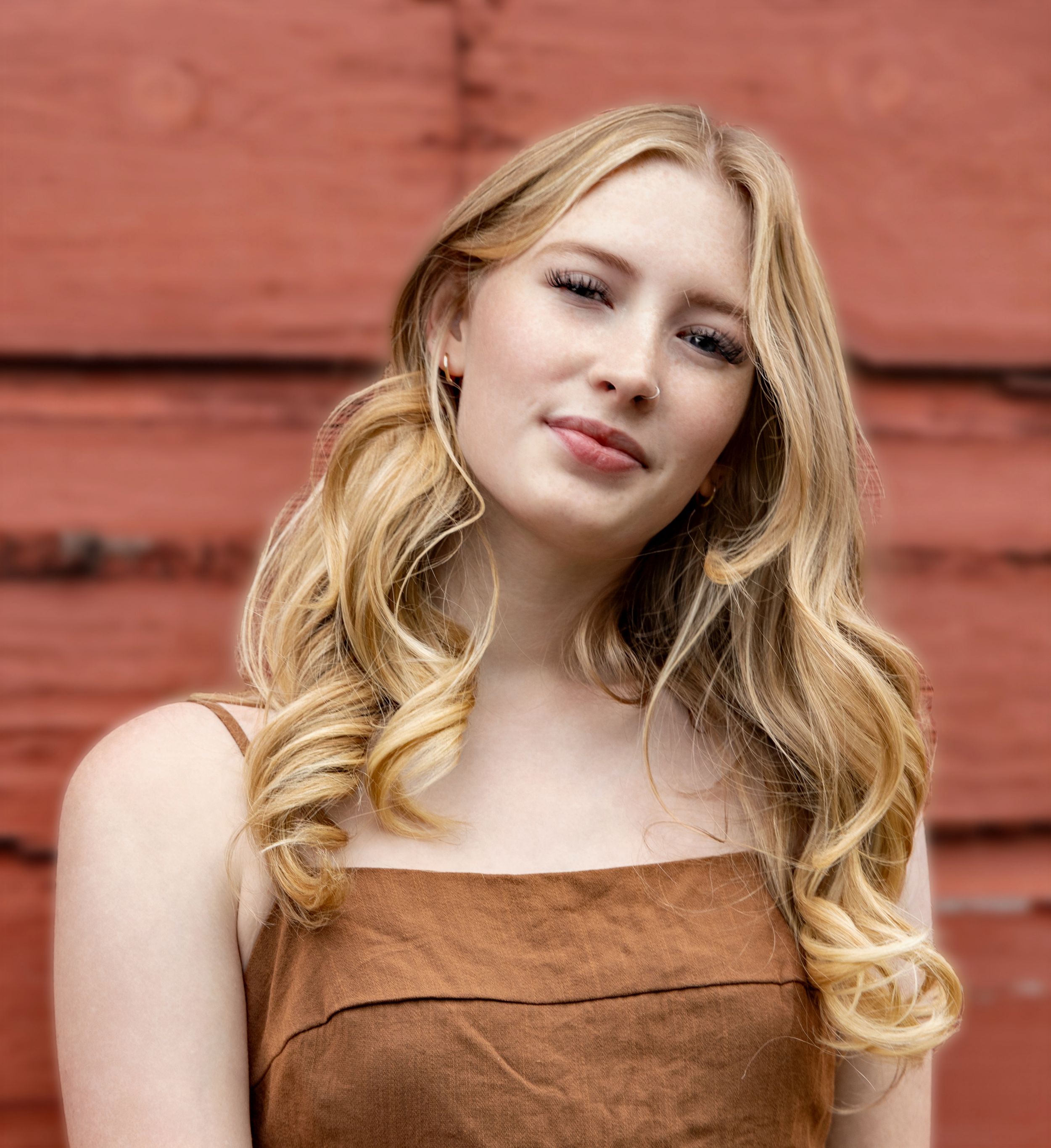 A person with blonde wavy hair poses against a rustic red barn wall in a brown sleeveless top.