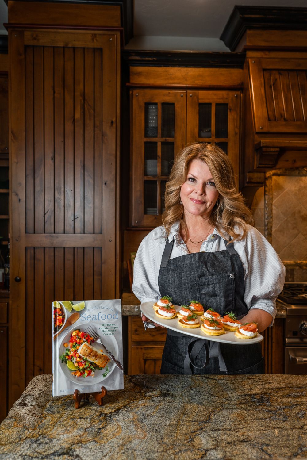 A chef in a gray apron holds a platter of fresh baked goods in a rustic kitchen with wooden cabinetry.